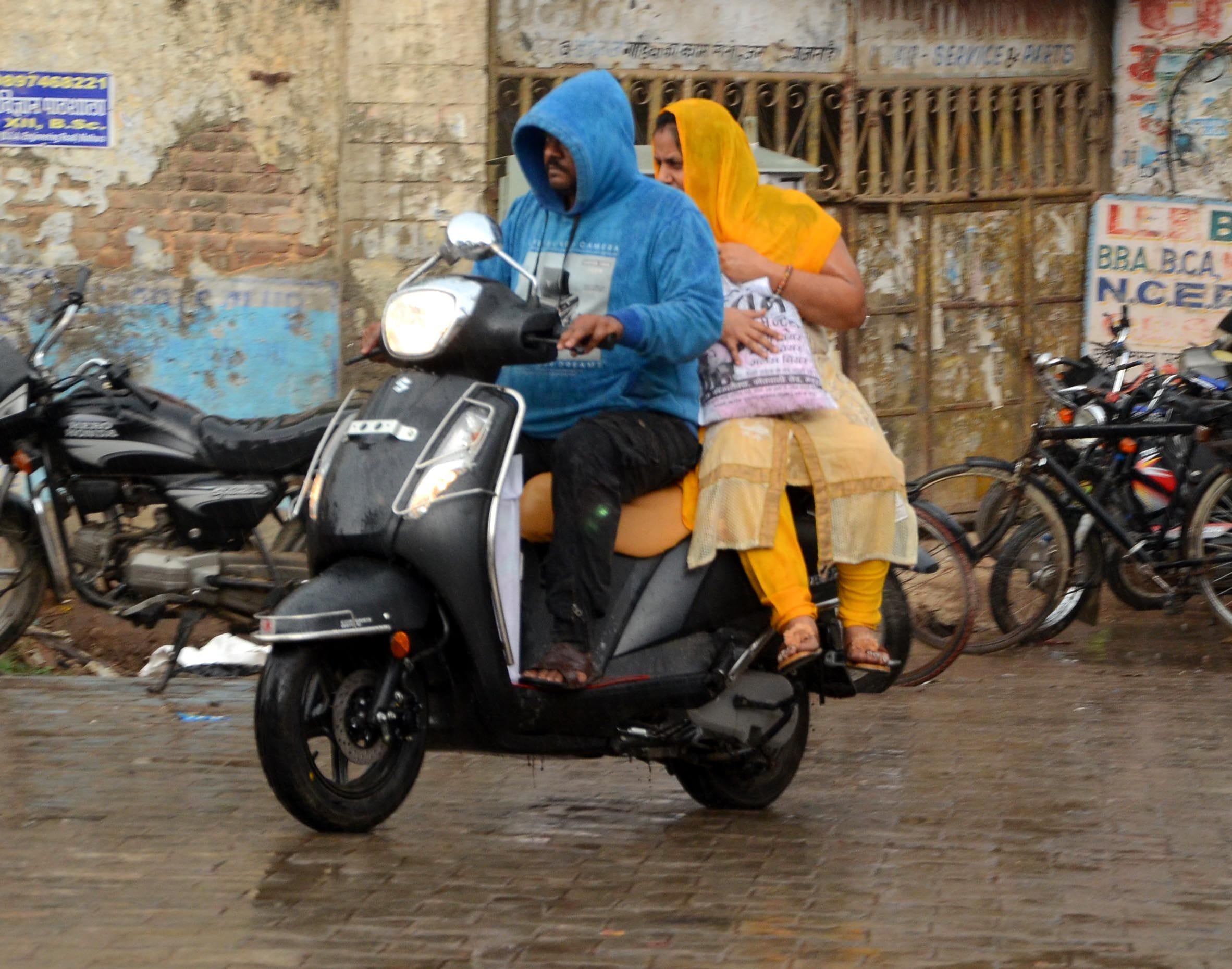 फोटो 51आर मौसम में आये बदलाव से हुई बारिश में भीगते जाते स्कूटी पर जाते दंपत्ति।