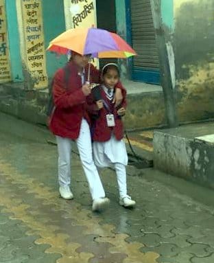 ऊन बरसात में छाता लगा कर जाते स्कूली बच्चे