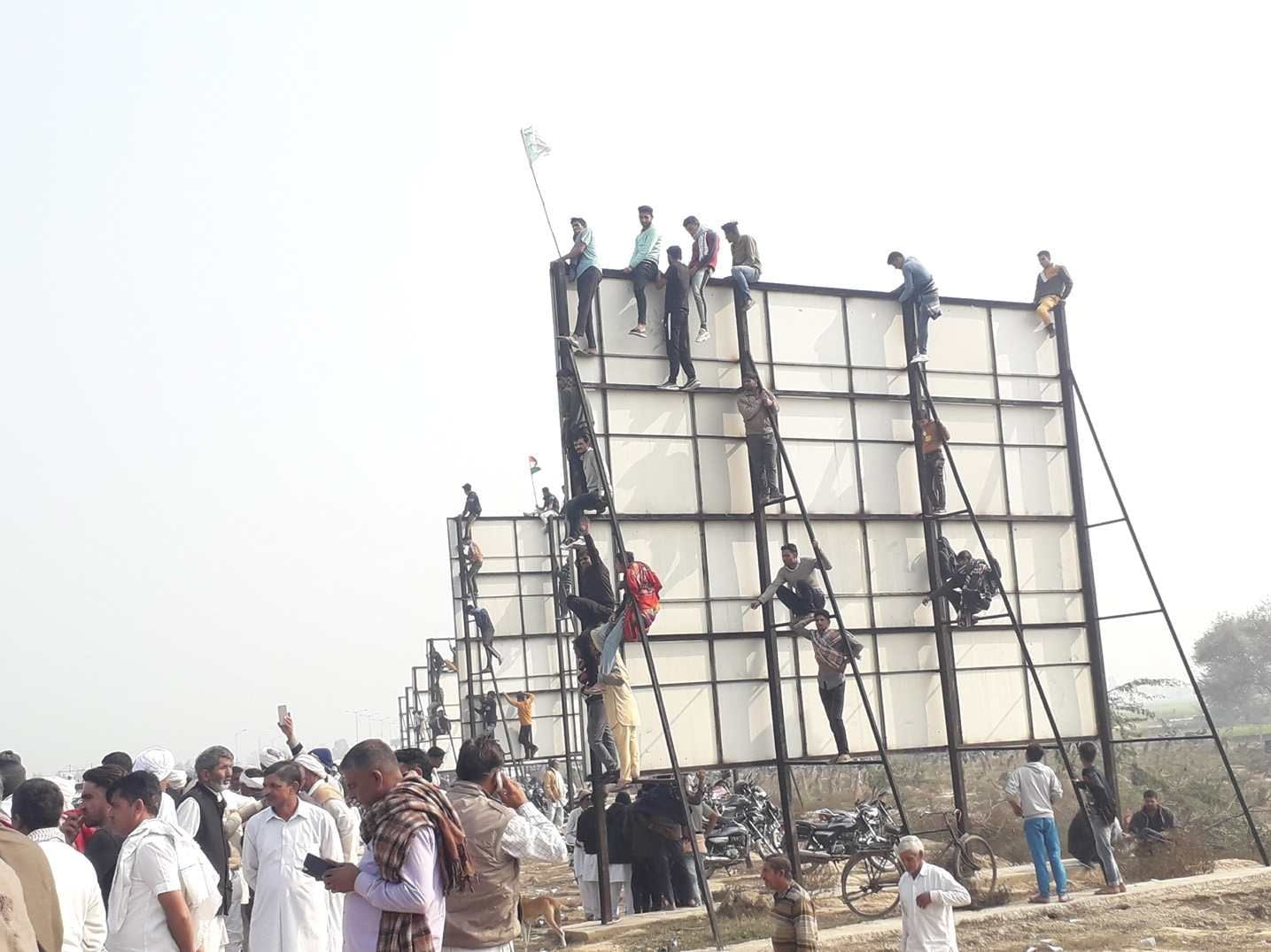 टोल पर लगे होर्डिंग्स पर चढ़कर राकेश टिकैत को देखते युवा। संवाद टोल पर लगे होर्डिंग्स पर चढ़कर राकेश टिकैत को देखते युवा। संवाद