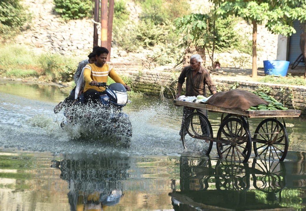 गंदे पानी से निकलता बाइक सवार, भीगने से बचता सब्जी विक्रेता। संवाद गंदे पानी से निकलता बाइक सवार, भीगने से बचता सब्जी विक्रेता। संवाद