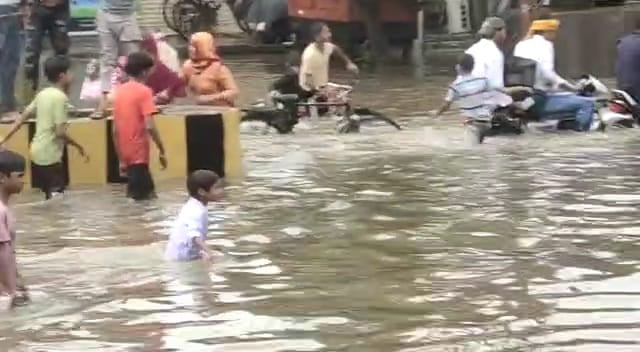 सड़क पर भरे पानी से गुजरते लोग और मस्ती करते बच्चें सड़क पर भरे पानी से गुजरते लोग और मस्ती करते बच्चें