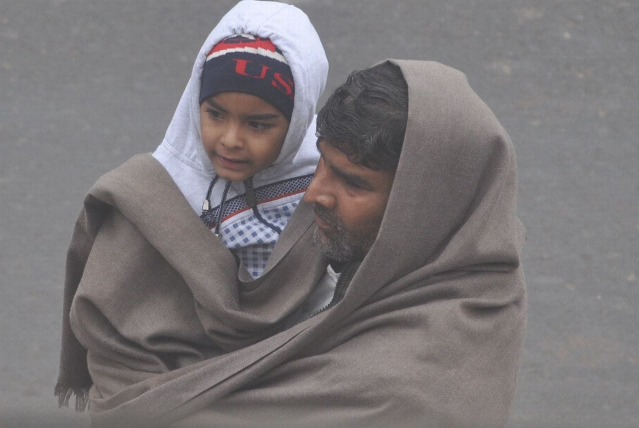 ठंड में बच्चे को गर्म चादर से लपेटकर गोद में ले जाता व्यक्ति । संवाद