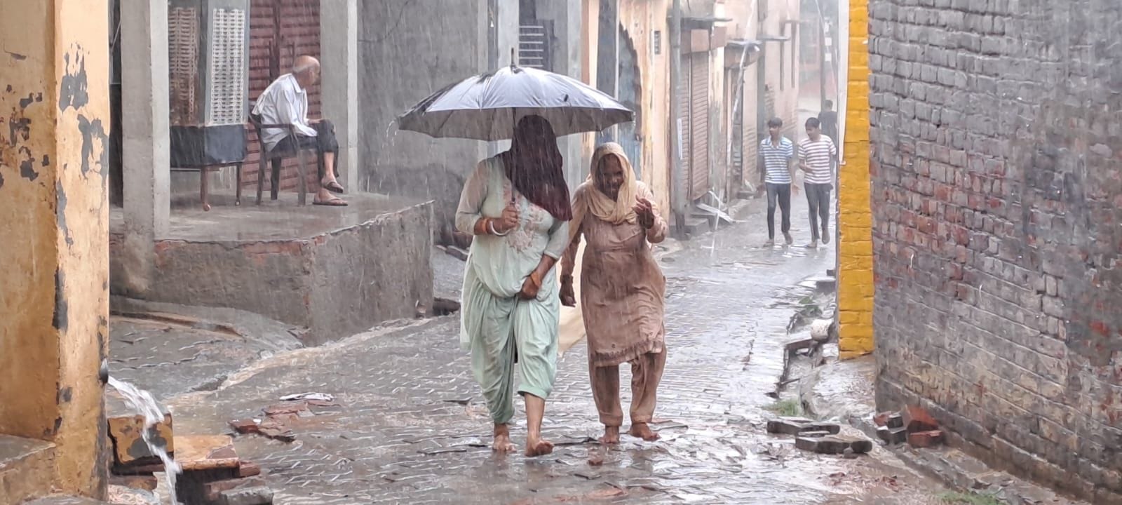 11जेएनडी37 : गांव मोरखी में हो रही बारिश के बीच से निकलती महिलाएं। संवाद