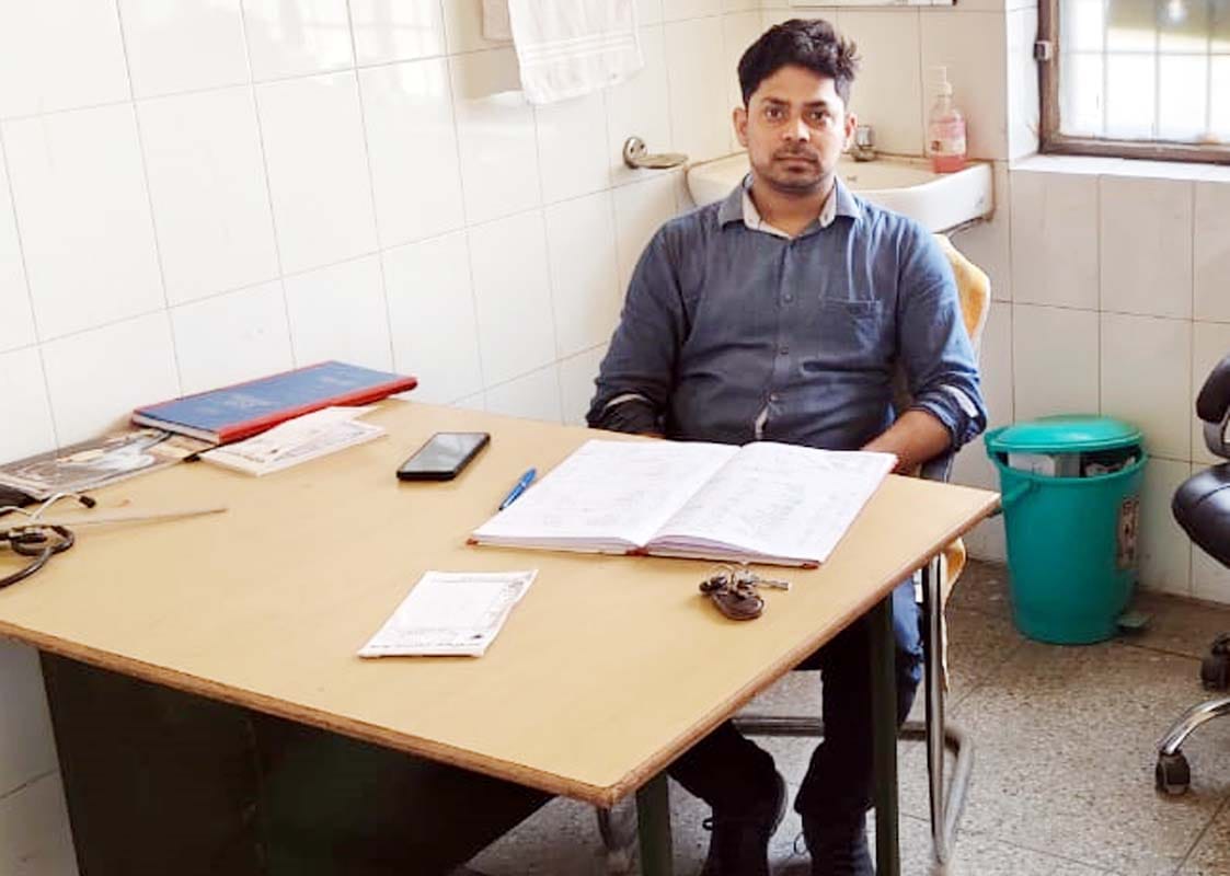नाहिल पीएचसी पर मेज पर पैर रखकर बैठा किशोर और रोगी देखते फार्मासिस्ट। संवाद